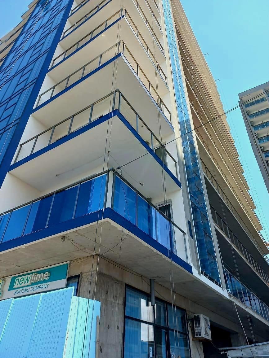 Реальная фотография фасада многоэтажного здания с нижнего ракурса, демонстрирующая сине-белую отделку, застекленные балконы и вывеску строительной компании.