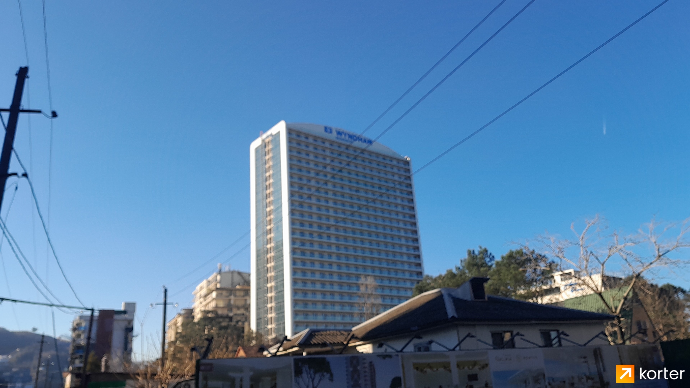 Реальная фотография экстерьера высотного здания отеля Wyndham на фоне голубого неба, с видными проводами и крышами соседних домов на переднем плане.